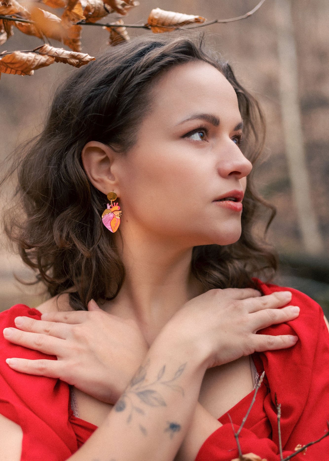 A woman with wavy brown hair and a tattooed forearm crosses her hands over her chest. She wears a red dress, bold style pink statement earrings, and stands outdoors among brown autumn leaves, looking to the side.