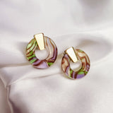 A pair of minimalist circle stud earrings featuring gold geometric tops and marbled, multicolored acrylic glass bases in pink, green, and beige tones, displayed on white satin fabric. Hypoallergenic for comfortable wear.