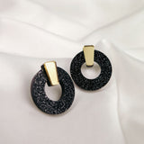 A pair of stylish Minimalist Circle Stud Earrings featuring black glittery acrylic glass hoops with gold rectangular accents, displayed on a white satin fabric background. Hypoallergenic for comfortable, all-day wear.