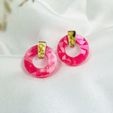 A pair of Minimalist Circle Stud Earrings with round, faceted pink acrylic glass and gold-tone accents rest on a smooth white fabric background. Delicate, out-of-focus dried flowers appear in the top left corner.