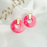 A pair of minimalist circle stud earrings with faceted pink acrylic glass and subtle gold accents, displayed on white satin fabric with soft, blurred dried plant accents in the foreground.