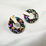 A pair of Minimalist Circle Stud Earrings made from hypoallergenic acrylic glass, featuring a black base with colorful, iridescent shards and gold-toned rectangular studs, displayed on a white fabric background.