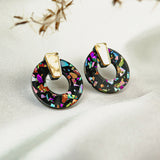 Close-up of Minimalist Circle Stud Earrings with gold tops and black acrylic glass bases featuring colorful, iridescent mosaic fragments, displayed on a white fabric background with dried plant stems overhead. Hypoallergenic design.