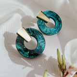 A pair of hypoallergenic, modern earrings with gold rectangular tops and circular blue-green marbled resin hoops, displayed on white fabric with a flower and buds nearby.