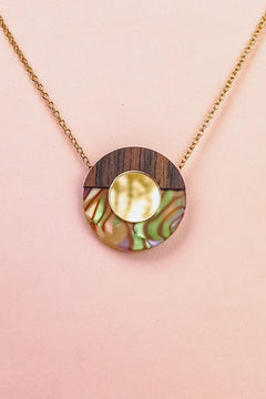 A minimalistic gold chain Circle Necklace with an oval pendant featuring wood, iridescent abalone shell, and a central circular gold detail, perfect for elevating any capsule wardrobe. Displayed against a pale pink background.
