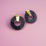 A pair of minimalist circle stud earrings, crafted from hypoallergenic acrylic glass with white speckles and gold rectangular accents, are displayed on a pink background.