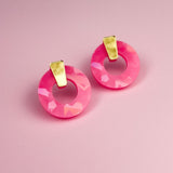 A pair of Minimalist Circle Stud Earrings featuring circular, faceted pink acrylic glass with elegant gold accents, displayed on a soft pink background.