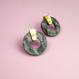 A pair of hypoallergenic Minimalist Circle Stud Earrings, featuring round green marbled acrylic glass and gold rectangular studs, displayed on a pink background.