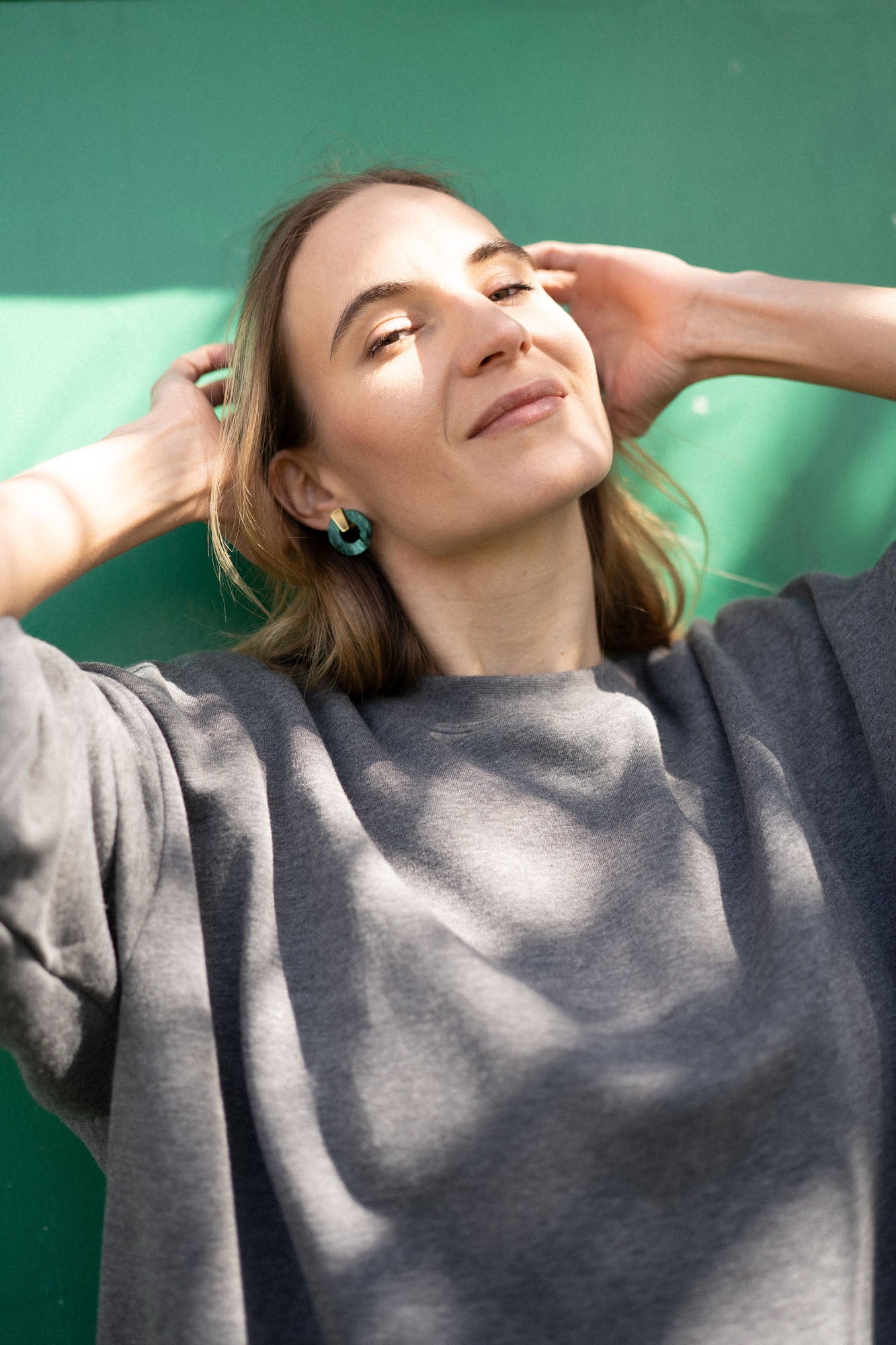 A woman with shoulder-length hair wearing a loose gray top stands in sunlight against a green background, smiling slightly with her hands raised near her head, showcasing Minimalist Circle Stud Earrings that are hypoallergenic.
