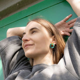A woman in a grey sweatshirt leans back with her arms raised behind her head, smiling slightly. She stands before a green wall, sunlight on her face, and wears hypoallergenic Minimalist Circle Stud Earrings in green acrylic glass.