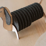 A napkin holder shaped like a dachshund dog, with a wooden head, legs, and tail, and black felt body segments holding napkins, sits on a light wooden table alongside matching dachshund coasters.
