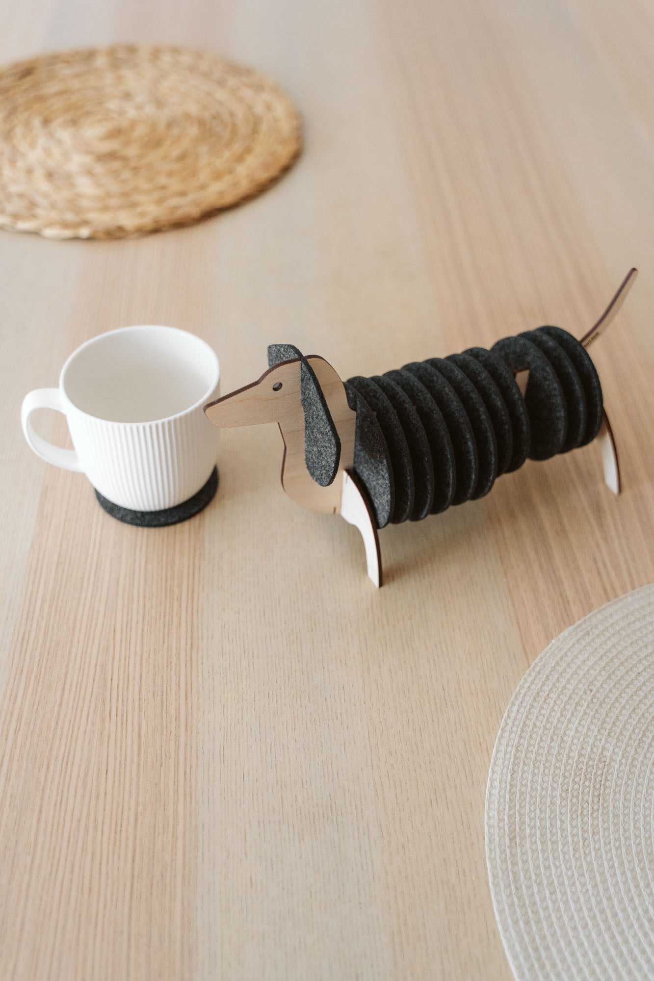 A wooden dachshund-shaped coaster holder with black round felt coasters on its body sits on a light wooden table next to a white mug, with woven placemats in the background.