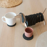 A dachshund-shaped coaster holder with black felt coasters on its body sits on a wooden table next to a white mug and a pink cup, both placed on coasters. Woven placemats are also visible on the table.