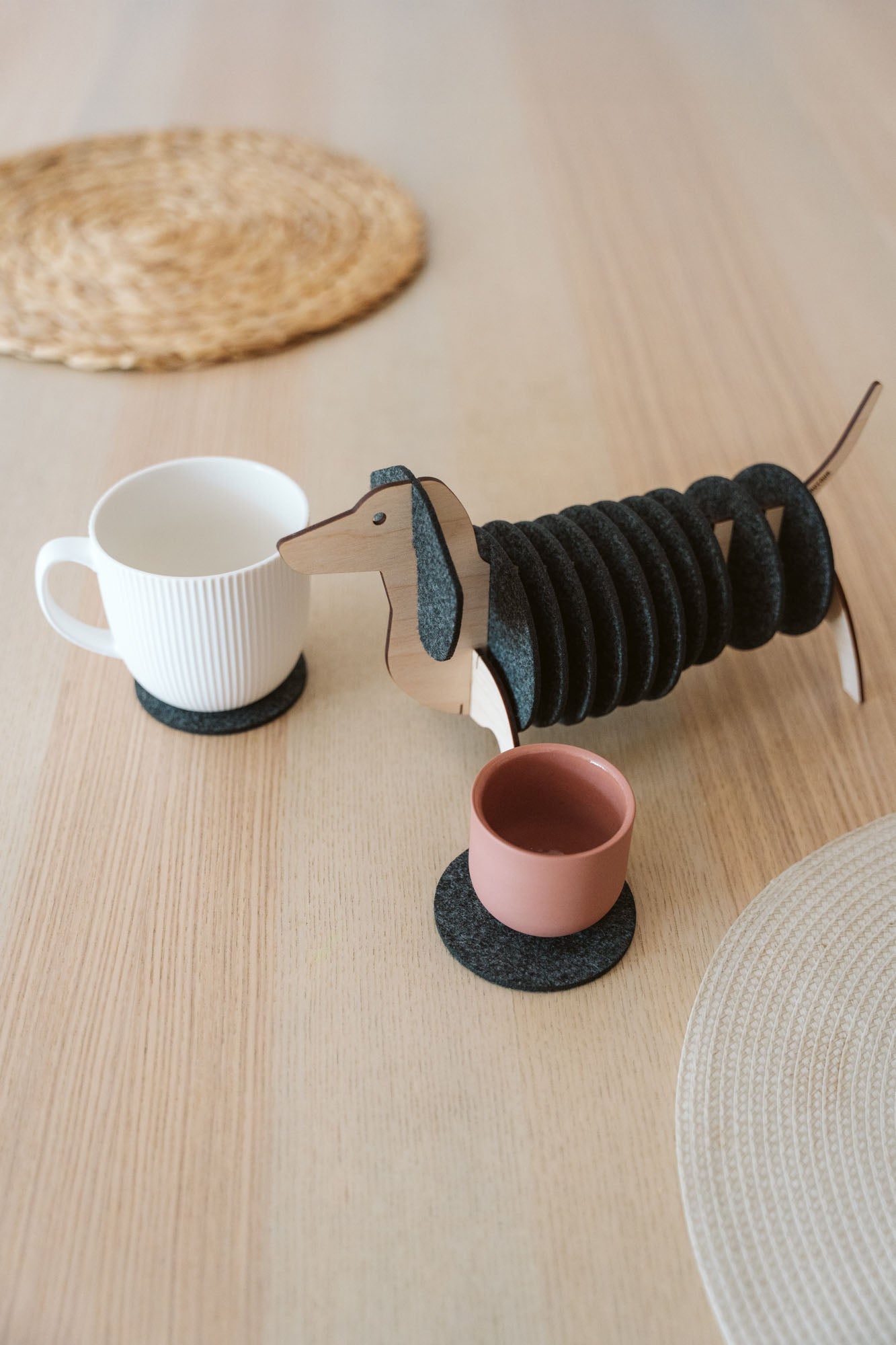 A dachshund-shaped coaster holder with black felt coasters on its body sits on a wooden table next to a white mug and a pink cup, both placed on coasters. Woven placemats are also visible on the table.
