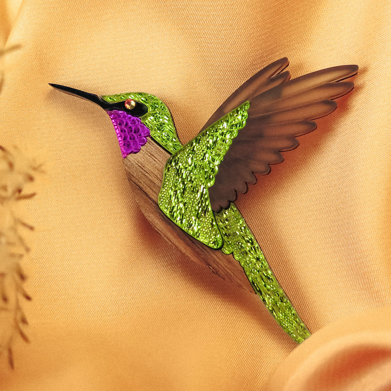 A handcrafted hummingbird brooch with vibrant green and purple glittery hues and brown wings rests on soft, peach-colored fabric.