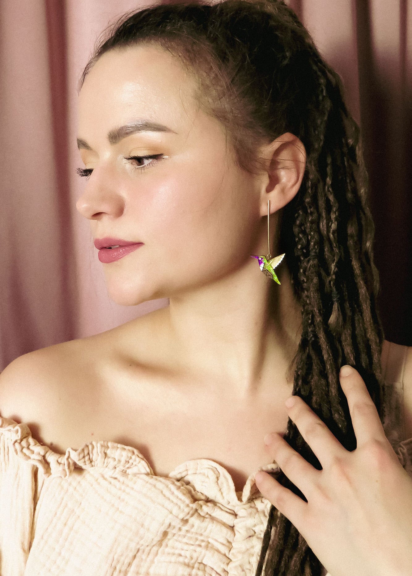 A woman with long brown dreadlocks and natural makeup wears an off-shoulder beige top, her hand to her chest. She showcases a colorful handcrafted hummingbird earring and stands before pink curtains, gazing thoughtfully to the side.
