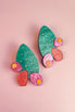 A pair of statement earrings shaped like green opuncia cactus pads with pink and orange flowers, decorated with small yellow dots, displayed on a light pink background.