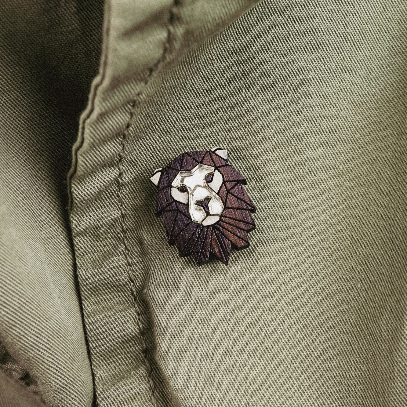 A close-up of an olive green jacket with a handcrafted geometric accessory—a Lion Pin—attached near a button.