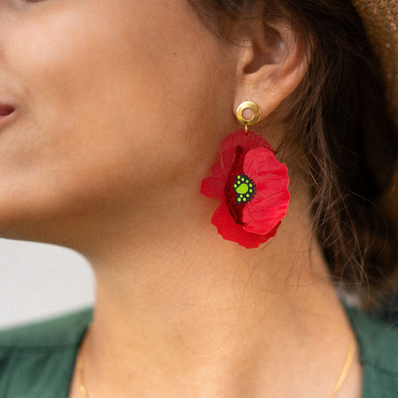 Close-up of a woman in a wide-brimmed hat, green top, and delicate gold necklace, wearing handcrafted Red Poppy Statement Earrings—a bold accessory with a vibrant red flower shape and green and black center. Her hair is tied back, and she smiles slightly.
