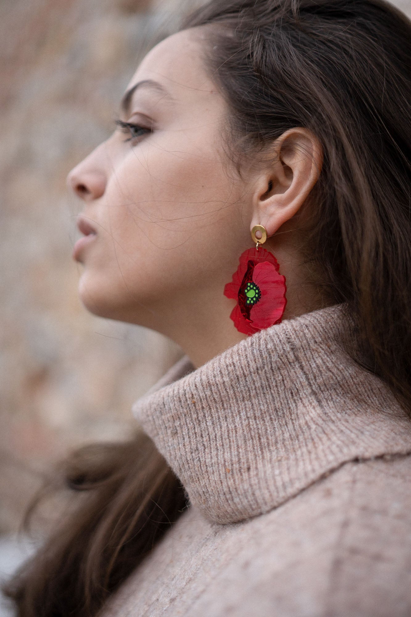 A woman wearing a beige turtleneck sweater is shown in profile. She has long brown hair and wears handcrafted Red Poppy Statement Earrings, a bold accessory with green and black details. The background is soft and out of focus.