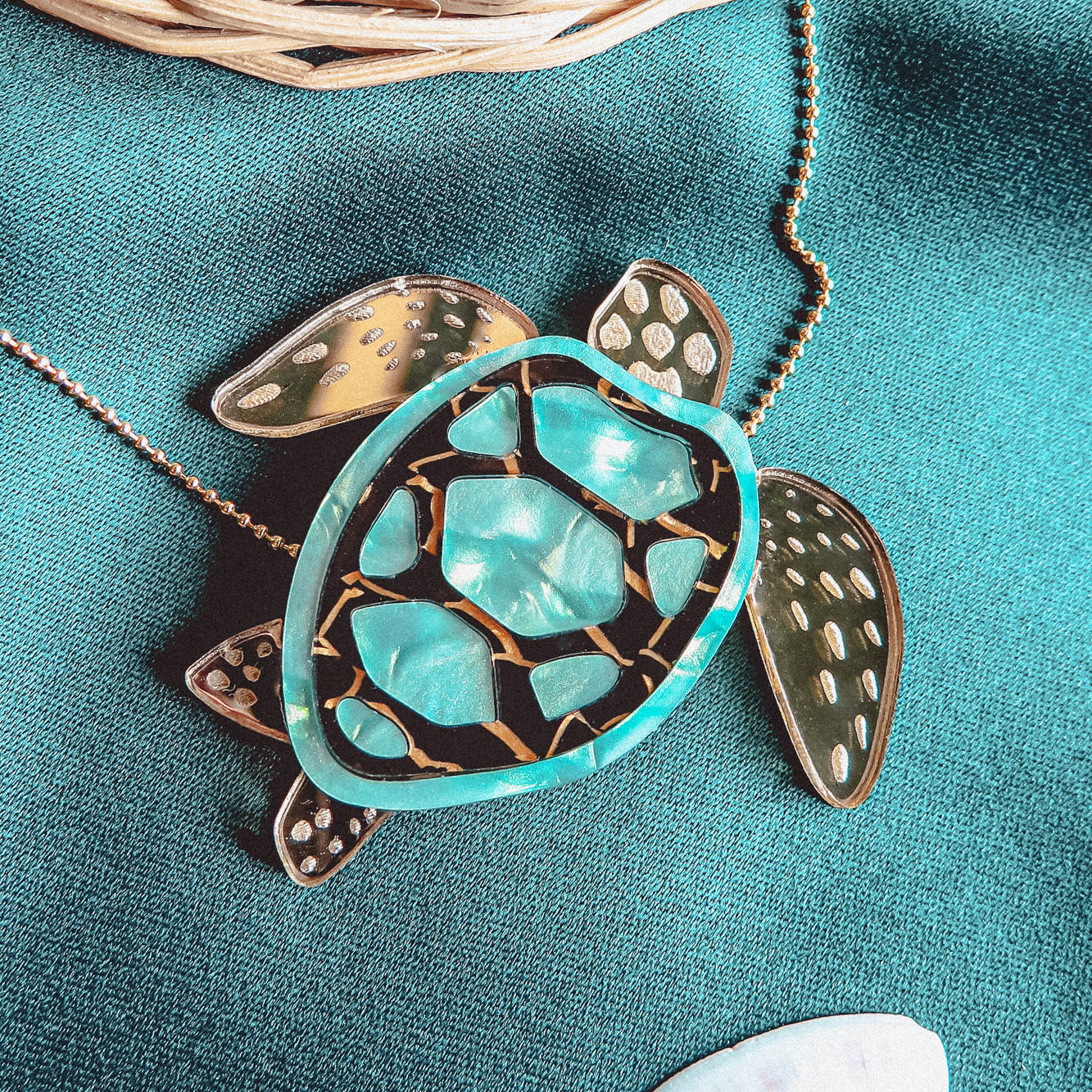 A close-up of a handcrafted Sea Turtle Necklace pendant with a turquoise and gold shell, lying on green fabric next to a white seashell and a wicker basket.