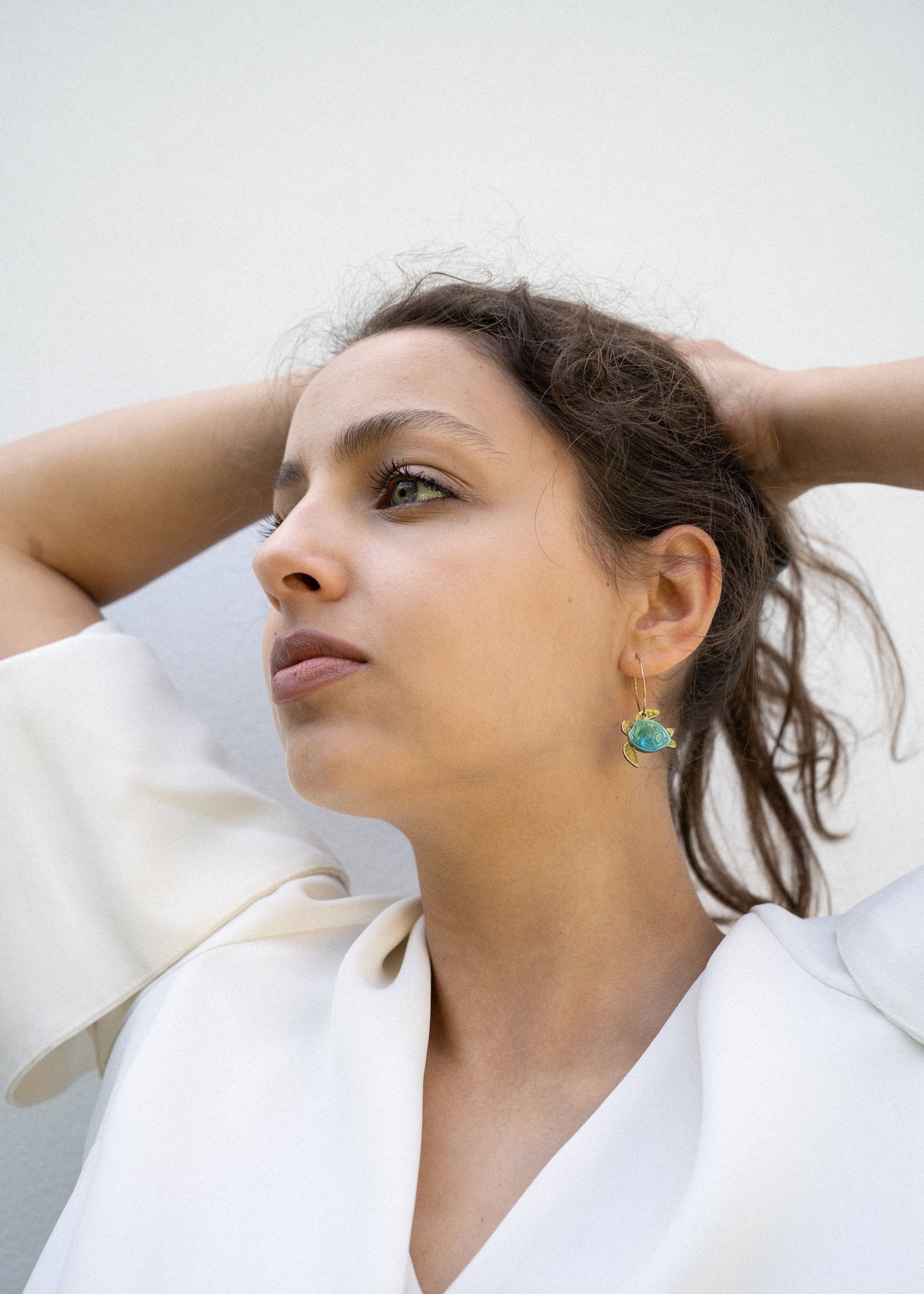 A woman with dark hair pulled back holds her hands behind her head, wearing a white blouse and handcrafted Little Turtle Hoop Earrings with green gemstones, looking thoughtfully into the distance against a plain light background.