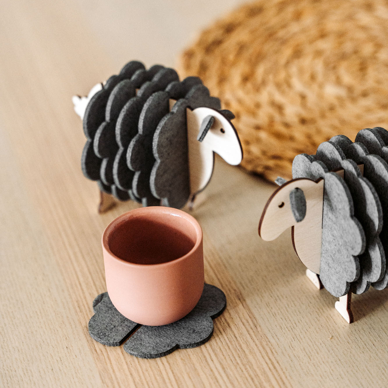 A pink cup rests on a gray, flower-shaped felt coaster beside two wooden sheep figurines—charming home decor—placed on a light wooden surface, with a woven mat partially visible in the background.