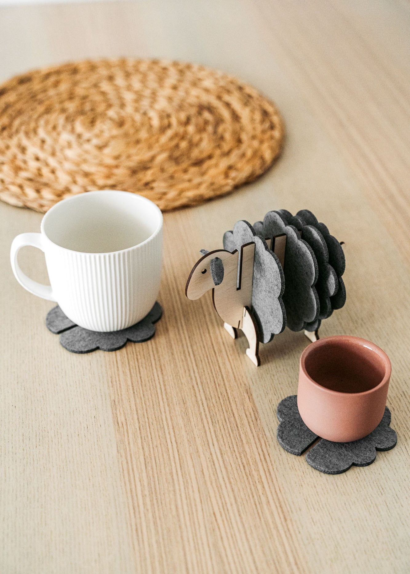 A white mug and a pink cup sit on dark felt coasters next to a decorative wooden sheep-shaped coaster holder, bringing charming home decor touches to a light wooden table topped with a round woven placemat.