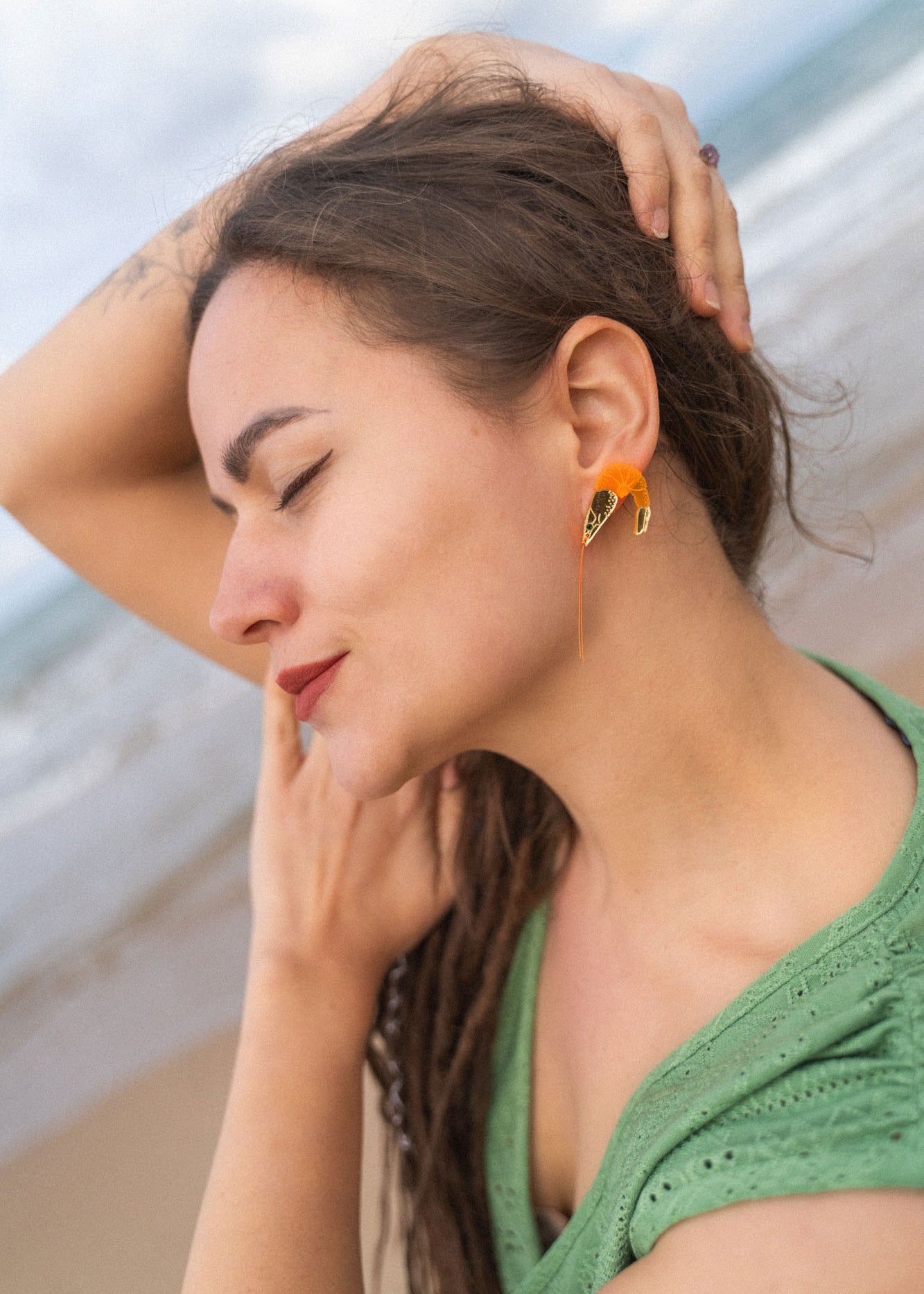 A woman with long brown hair and a green shirt stands on a beach with her eyes closed, smiling gently, and one arm raised over her head, showing off Shrimp Stud earrings sparkling with emerald rhinestones.