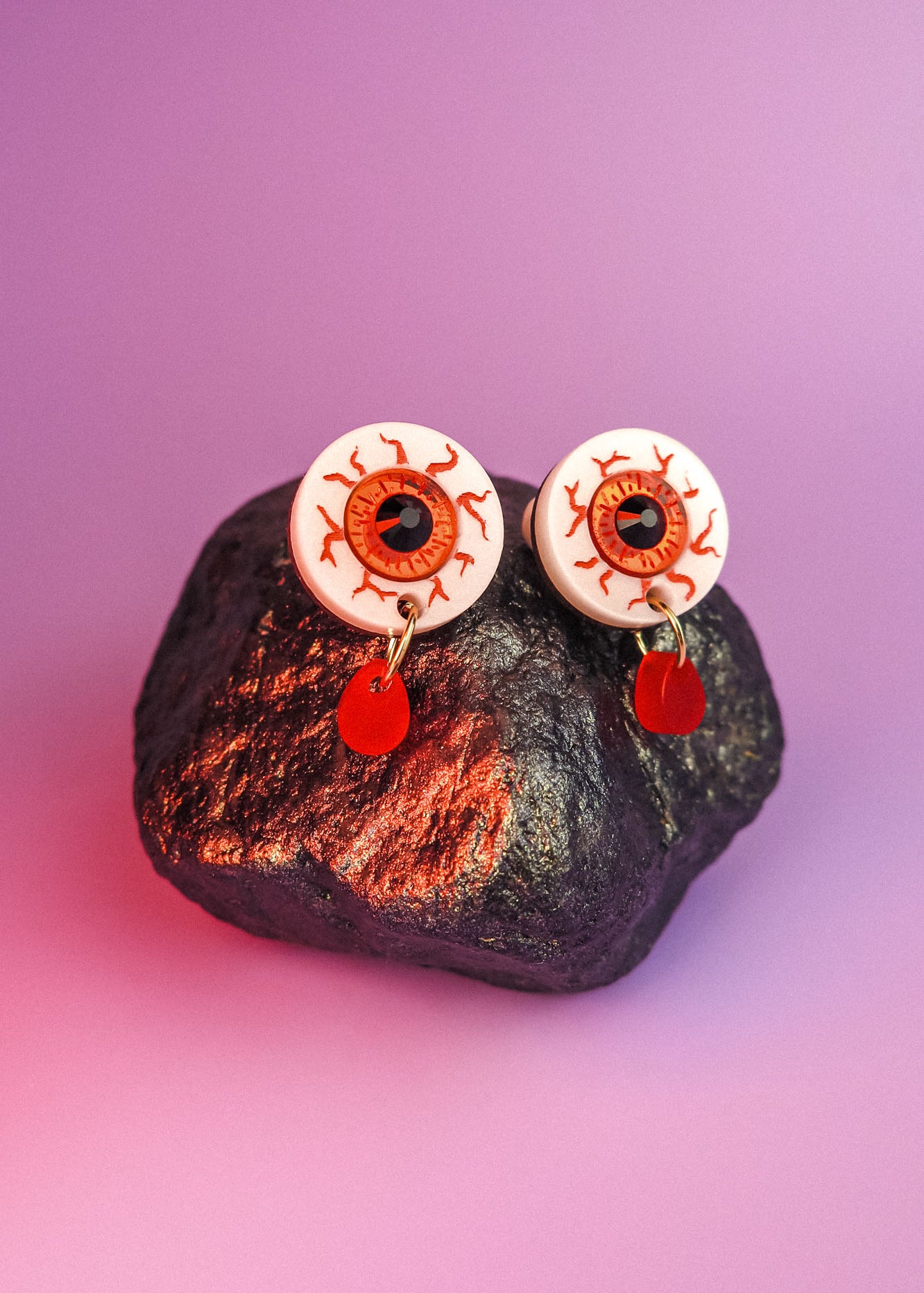 A pair of hand-painted earrings designed like bloodshot eyeballs with red teardrop-shaped pendants hang from a dark, textured rock against a purple background.
