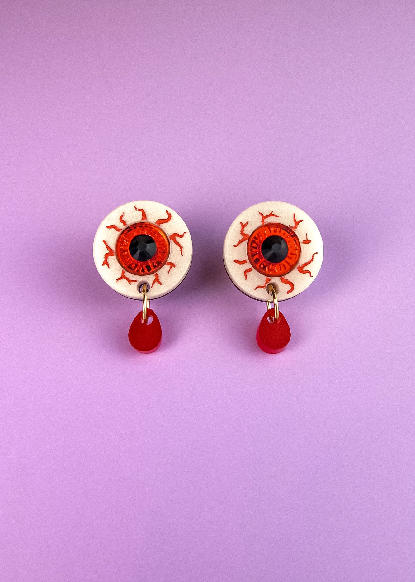 Two hand-painted earrings designed as white eyeballs with red veins and black pupils, each featuring a dangling red teardrop, are displayed on a light purple background. These allergy-free earrings add a quirky, artistic touch to any look.