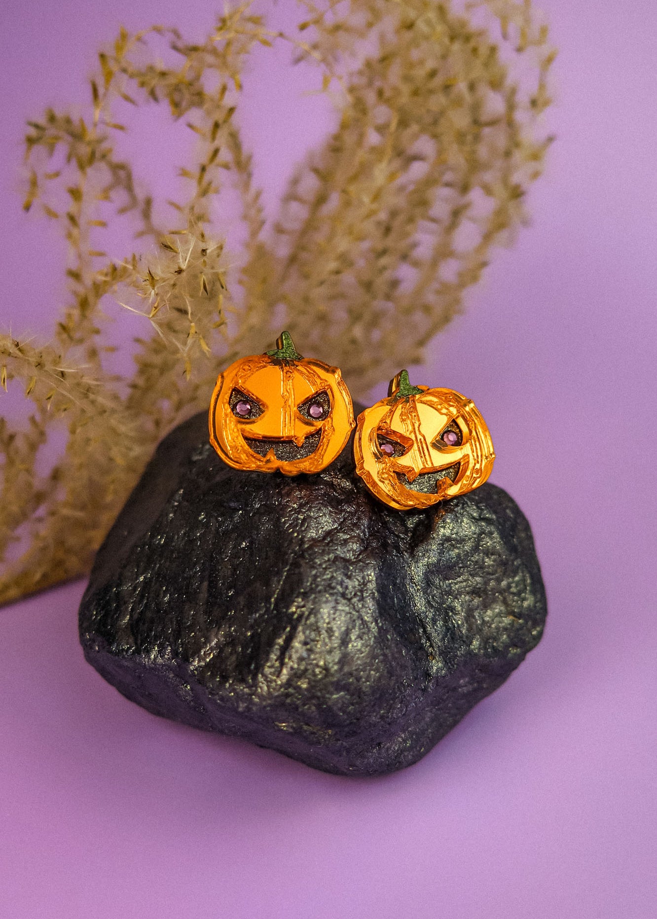 Two small spooky pumpkin studs with carved faces sit on a black rock, with dried grass in the background and a purple surface underneath—perfect Halloween earrings for fans of handcrafted jewelry.
