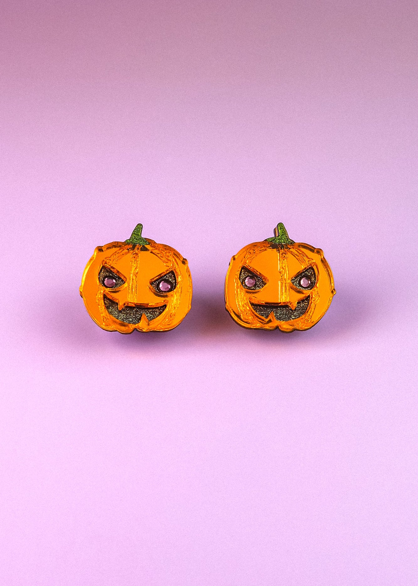 Two small jack-o-lanterns with angry expressions are placed side by side on a pink and purple gradient background, capturing the perfect mood for Spooky Pumpkin Studs—ideal handcrafted jewelry for Halloween earrings lovers.