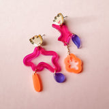 Two mismatched earrings in bold colors of pink, orange, and purple acrylic, featuring playful abstract shapes. Each unique earring dangles with geometric forms, making these Mismatched Earrings a vibrant statement on a light pink background.