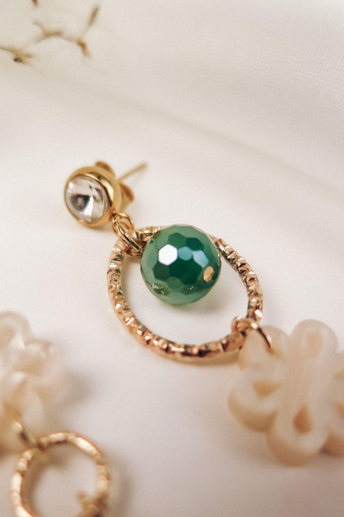A close-up of a gold earring featuring a small clear stone, a textured circular gold loop, a faceted green bead, and an off-white decorative charm, blending modern charm with timeless elegance against a soft, white background.