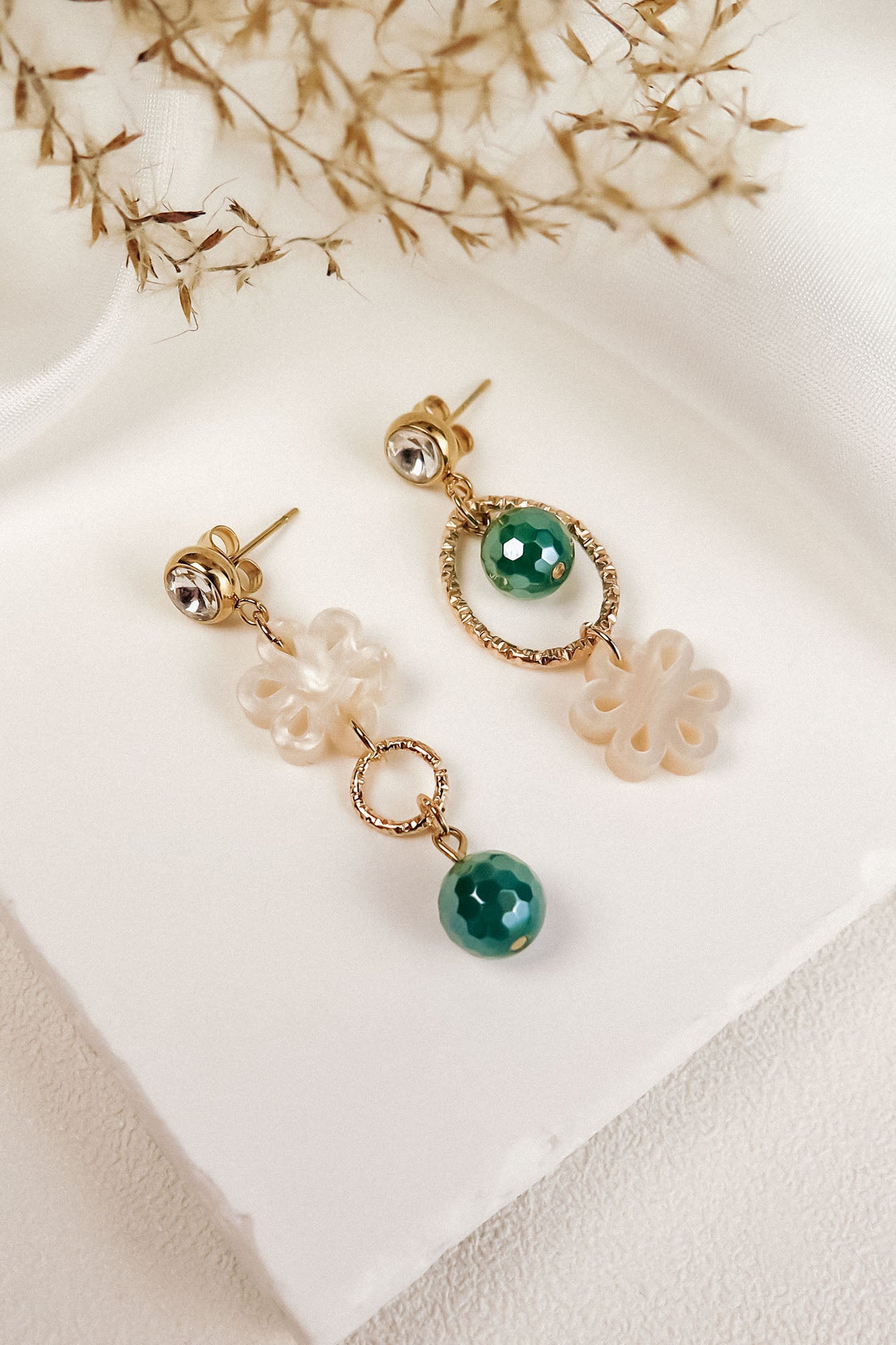 A pair of mismatched earrings displayed on a white surface, featuring green gemstones, gold accents, and white floral-shaped elements, exuding timeless elegance with delicate dried grass in the background.