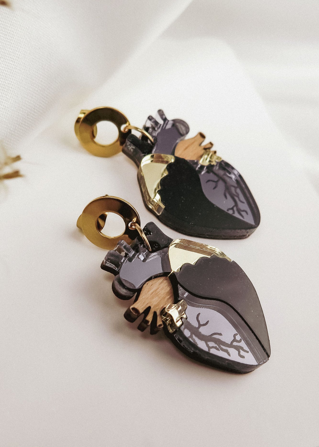 A pair of Anatomical Heart Earrings with gold accents and black, grey, and wood-toned details adds a touch of gothic flair, displayed on a white fabric background—perfect for lovers of unique handcrafted accessories.