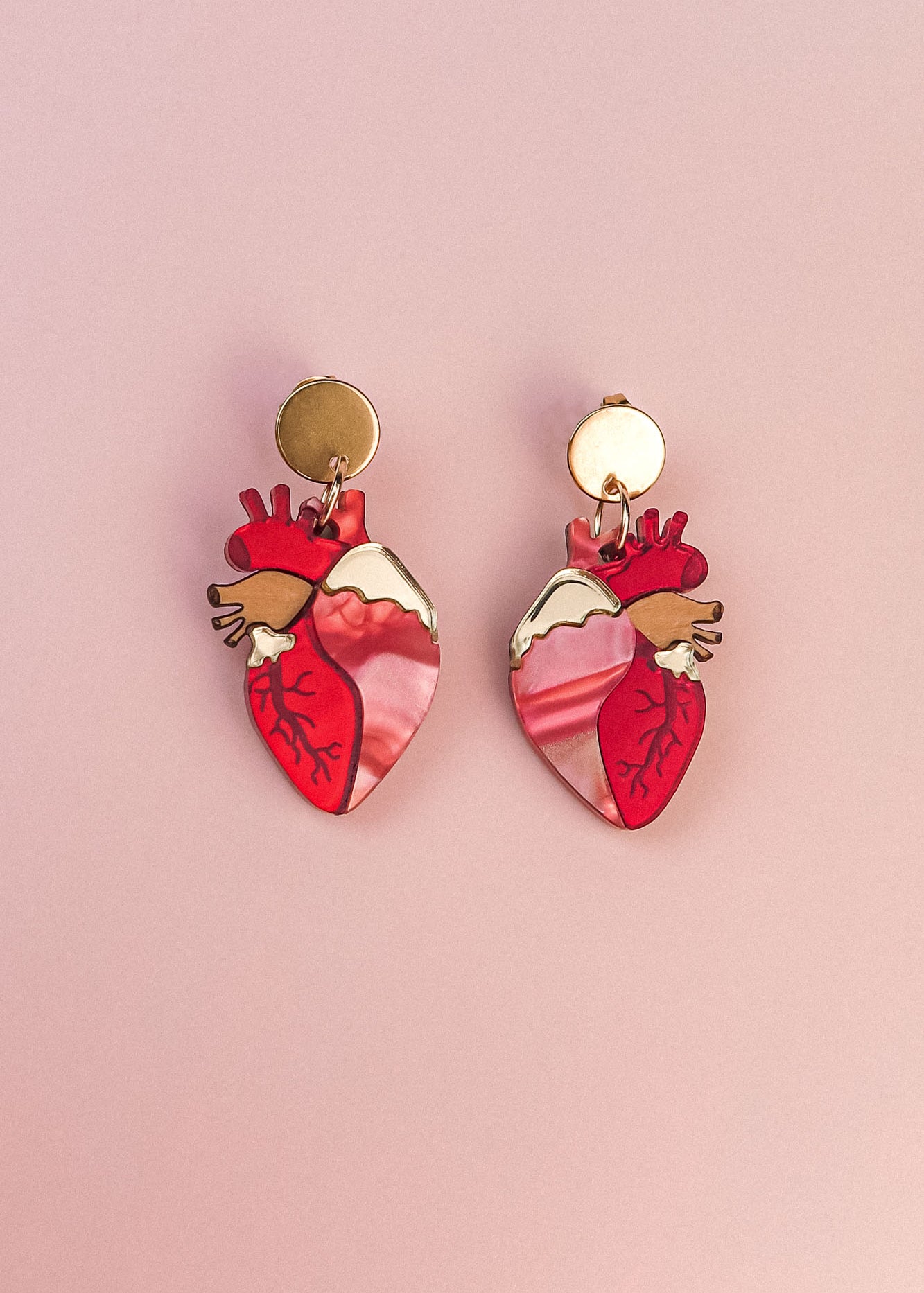 A pair of gold and pink Anatomical Heart Earrings, crafted in acrylic with detailed vein patterns and a unique style, displayed on a light pink background.