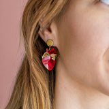 Close-up of a woman’s ear wearing unique style Anatomical Heart Earrings in red and pink tones with gold accents; her light brown hair is tucked behind her ear against a pink background, highlighting these striking acrylic earrings.