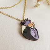 Gold chain necklace with a pendant shaped like an anatomical heart, featuring purple, black, gold, and wooden accents—this handcrafted jewelry piece is displayed on a light textured surface with dried grasses nearby.