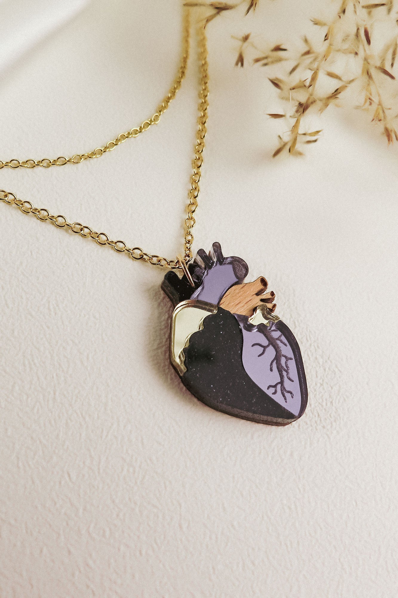 Gold chain necklace with a pendant shaped like an anatomical heart, featuring purple, black, gold, and wooden accents—this handcrafted jewelry piece is displayed on a light textured surface with dried grasses nearby.