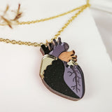 A gold chain necklace features a pendant shaped like an anatomical heart, with black, purple, and gold details—an eye-catching handcrafted jewelry piece resting on a white textured background.