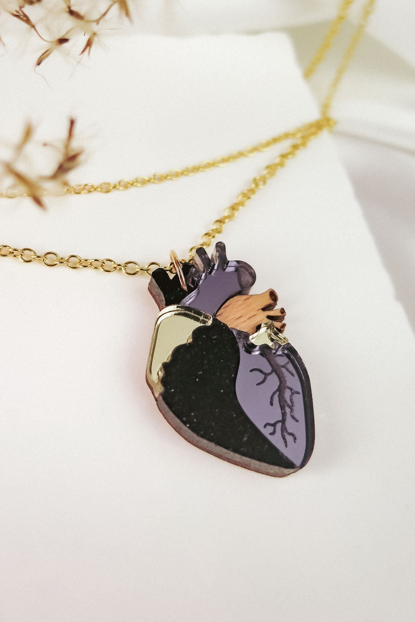 A gold chain necklace features a pendant shaped like an anatomical heart, with black, purple, and gold details—an eye-catching handcrafted jewelry piece resting on a white textured background.