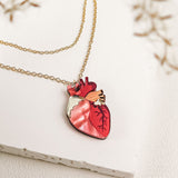 A gold chain anatomical heart necklace with a pendant shaped like a human heart, featuring red and pink detailing, is displayed on a white textured surface with delicate blurred foliage in the foreground—a truly unique jewelry piece.
