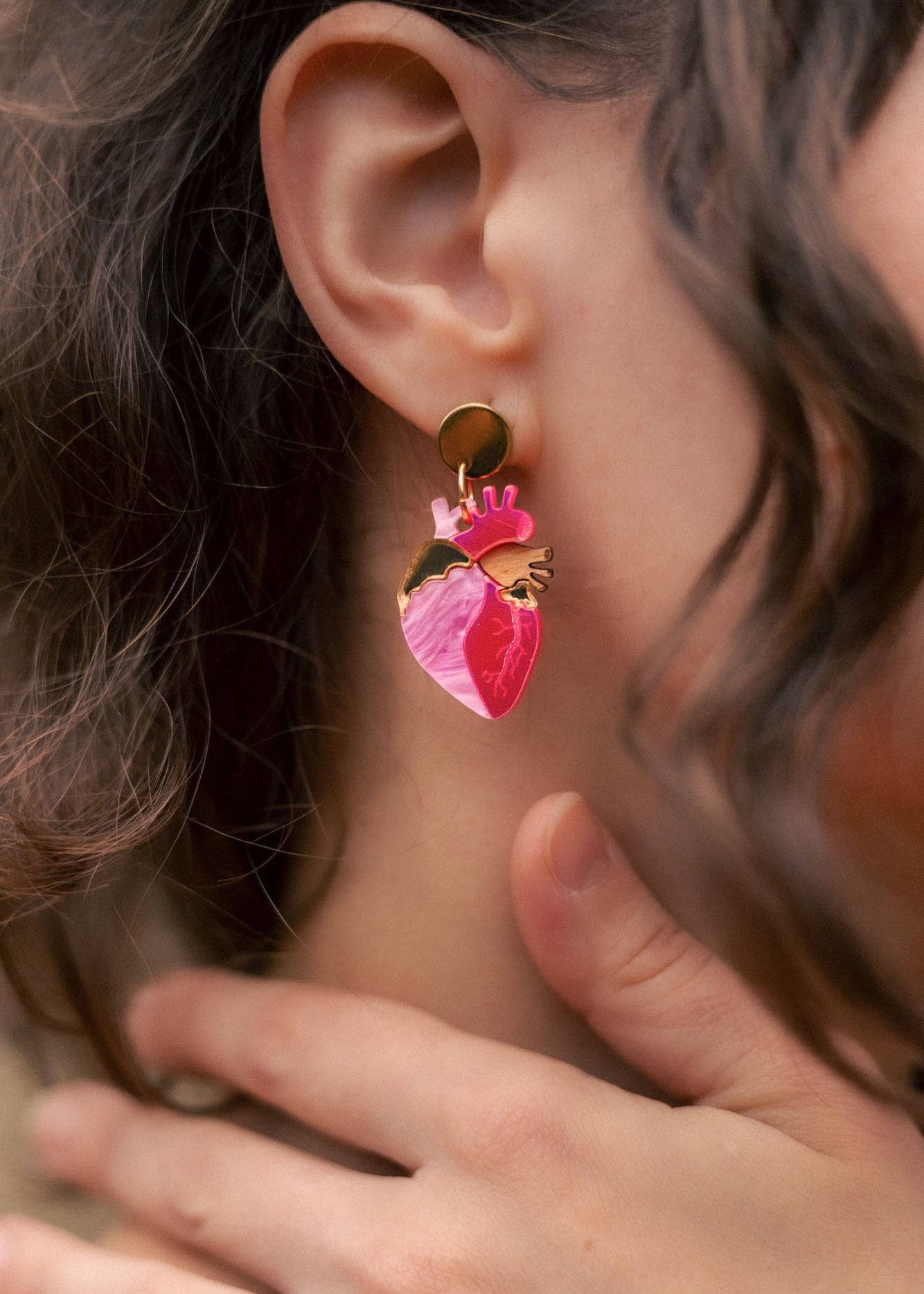 A close-up of a person’s ear wearing bold Anatomical Heart Earrings crafted from pink and gold holographic acrylic. Their curled brown hair partially frames the ear, while their hand gently touches their neck.