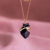 A handcrafted anatomical heart necklace with gold and dark purple details is captured against a soft pink textured background, highlighting this unique jewelry piece’s artistic design.