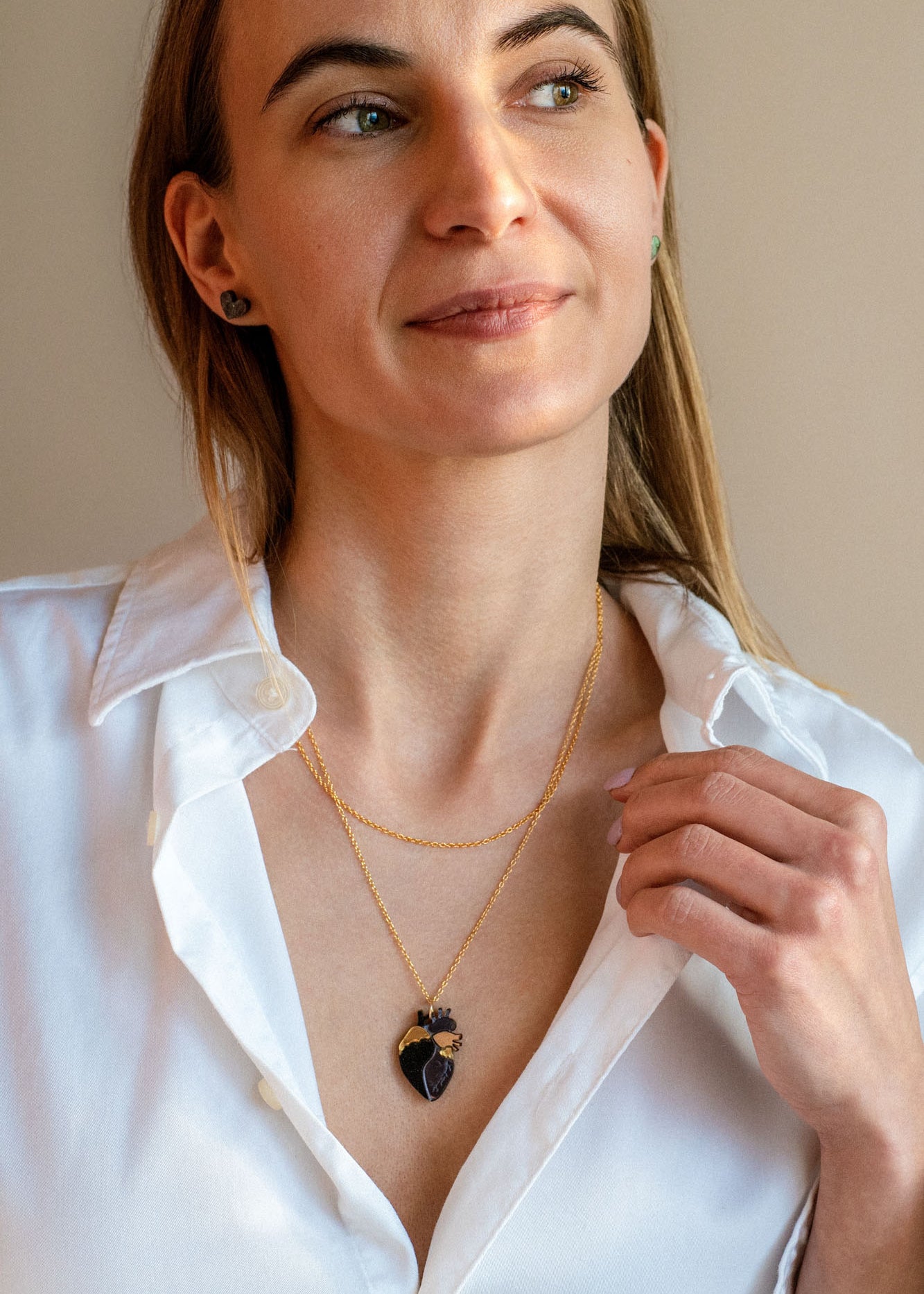 A woman with long light brown hair wears a white button-up shirt and a gold anatomical heart necklace, looking slightly to the side with a soft smile against a plain beige background.
