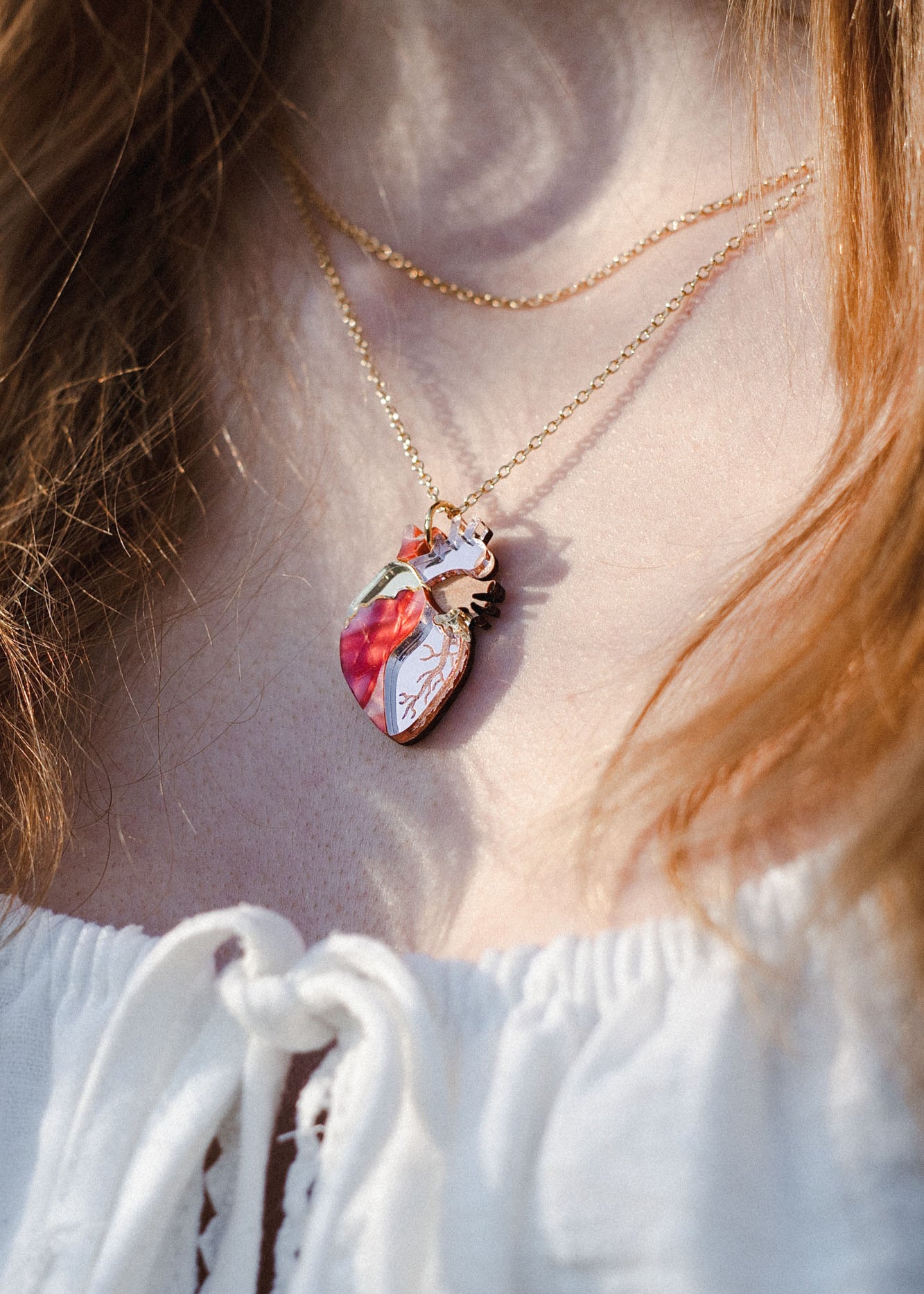 Close-up of a person wearing an anatomical heart necklace in red, pink, and white—a striking piece of unique jewelry. The handcrafted pendant hangs over a white top with a lace-up detail.
