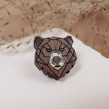 A handcrafted wooden brooch in geometric style, shaped like a bear’s head, rests on a white surface with delicate dried plants softly blurred in the background.
