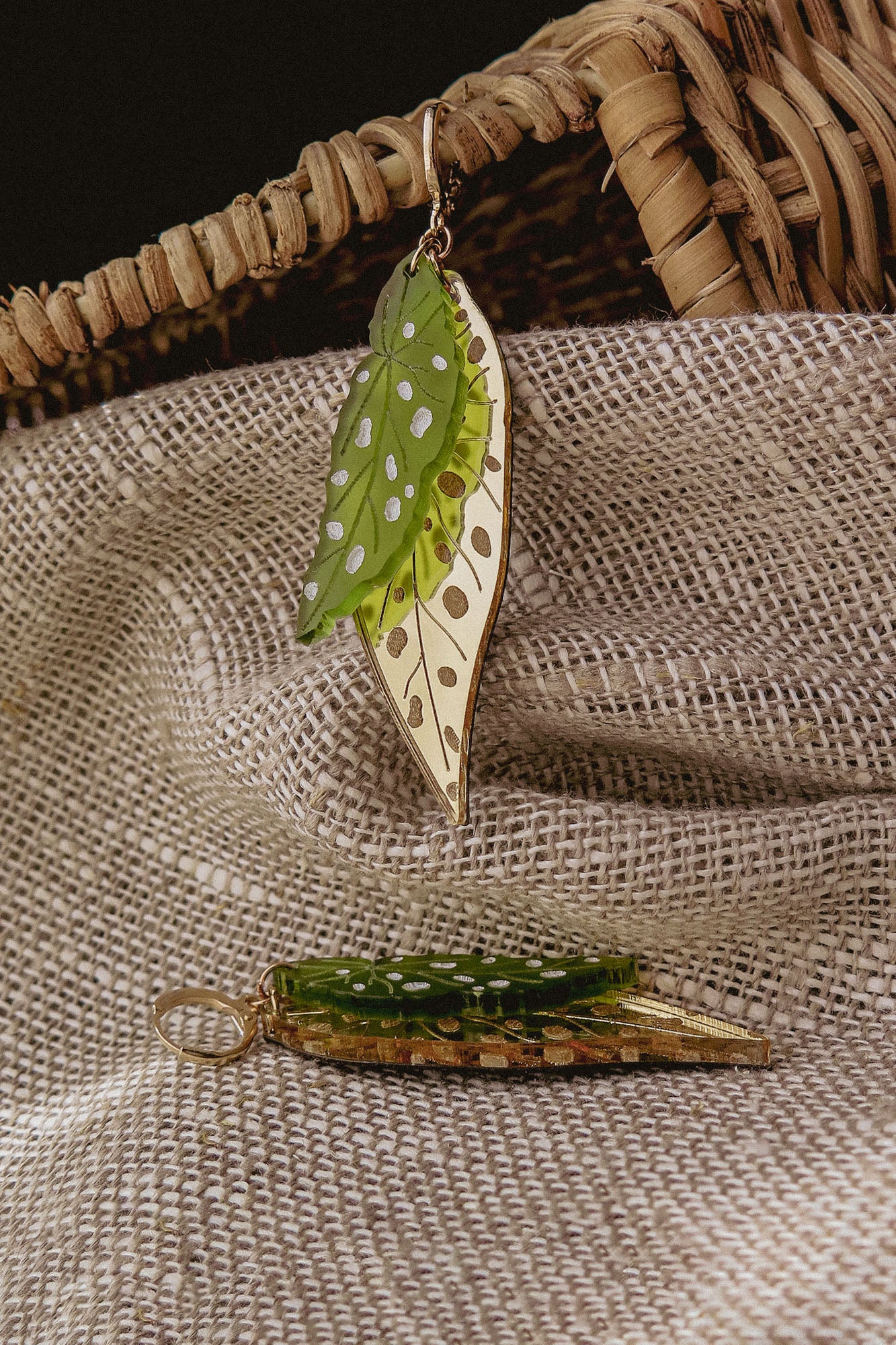 A pair of Begonia Leaves Earrings with green and gold colors and white spots rest on textured beige fabric near a woven basket, making them unique jewelry perfect for plant lovers.